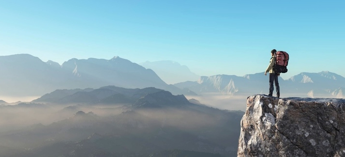 Hiker with a backpack on a cliff looking at mountains.